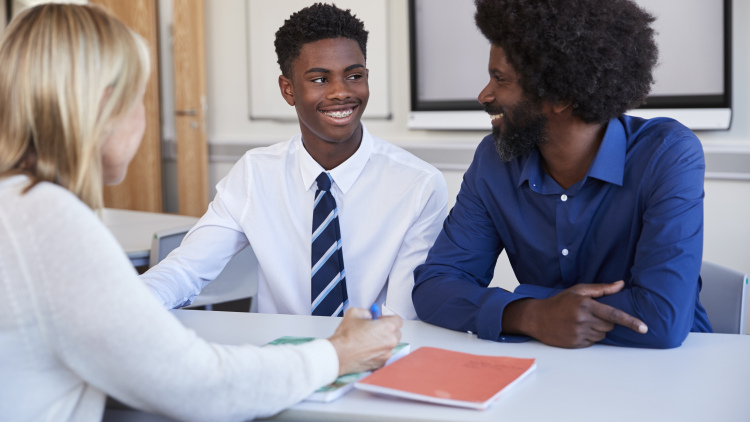 A black father and teenage son discuss with a female teacher at a high school parents' evening.